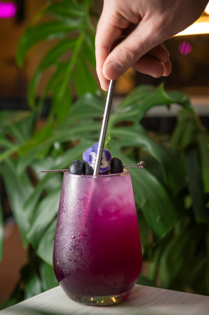 Coctel magenta servido con flores comestibles sobre fondo tropical.